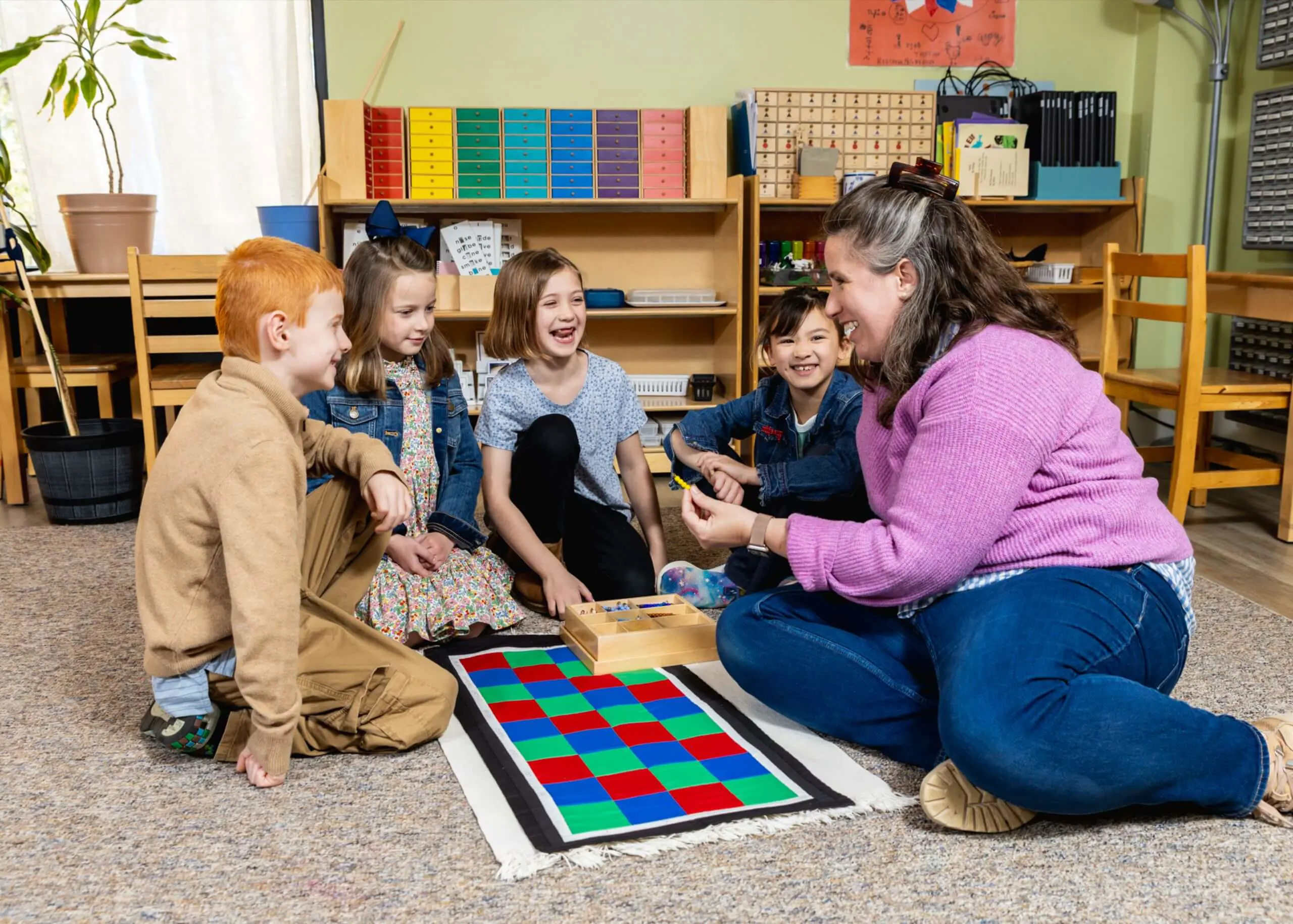 A teacher sitting on the floor with three elementary students working together on a Montessori checkerboard math activity.
