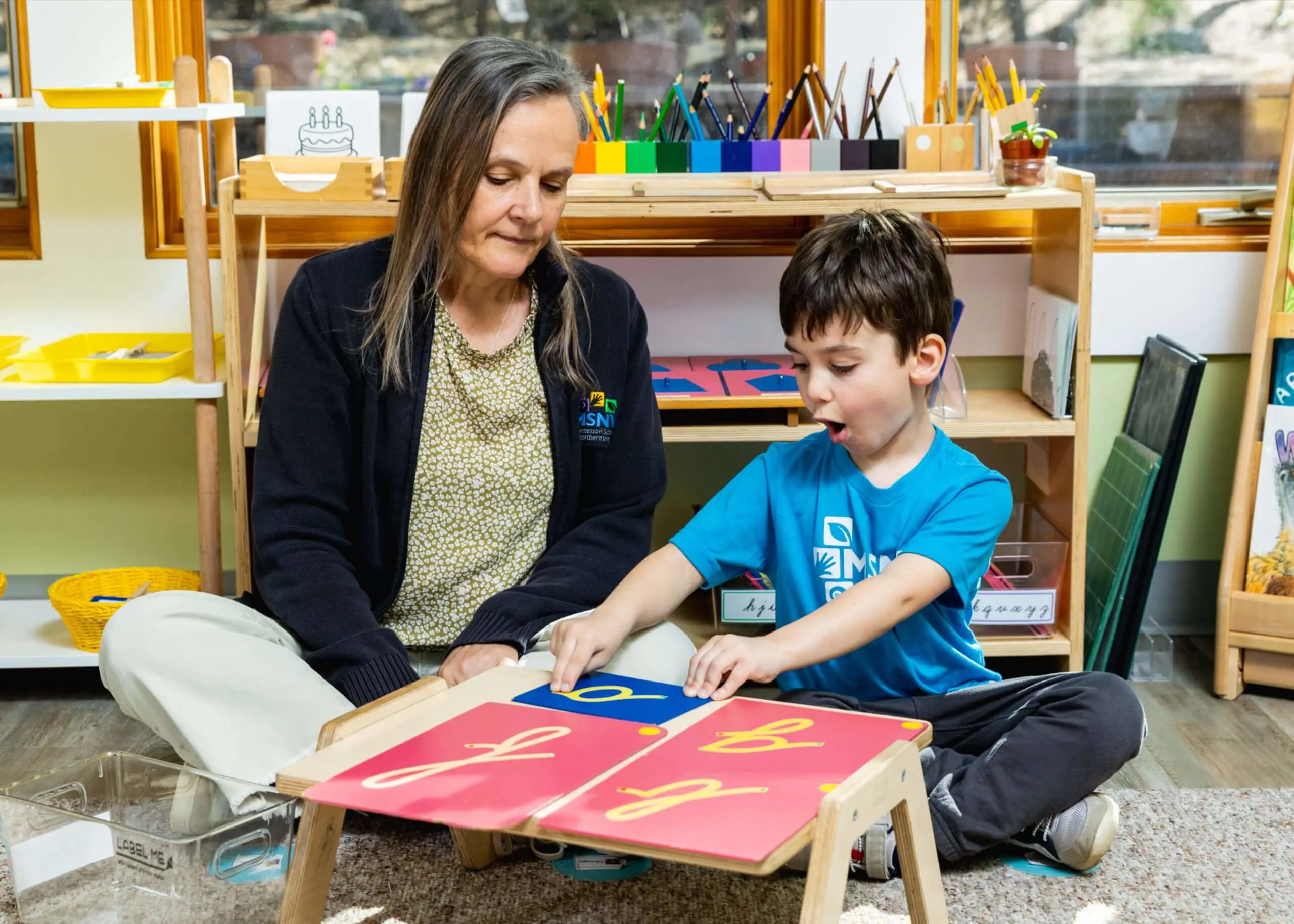An MSNV teacher sitting beside a primary-age boy and guiding him through a Montessori activity at a low table.