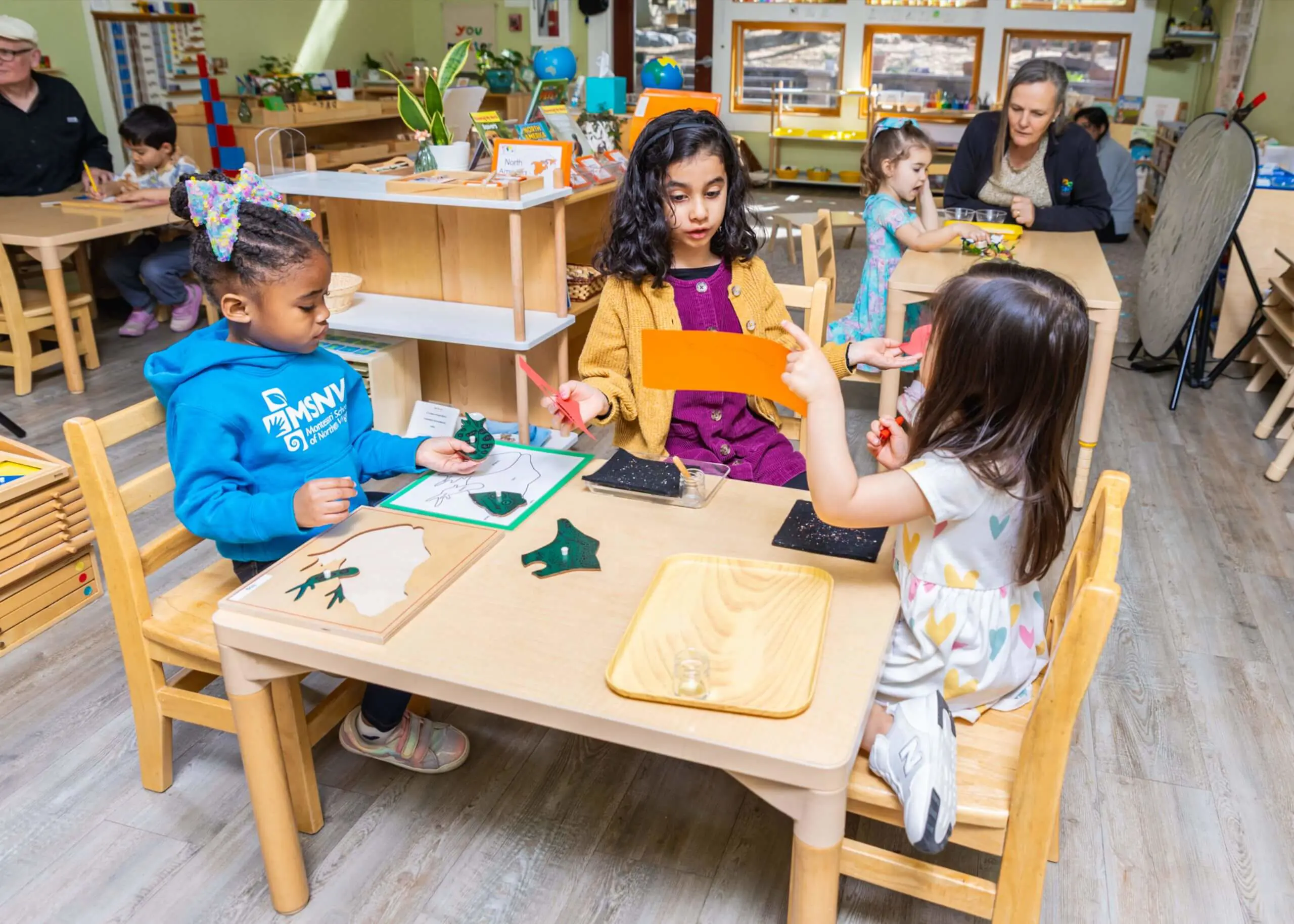 Three MSNV Primary students engaged in independent and small group work at a classroom table, with a teacher observing in the background.