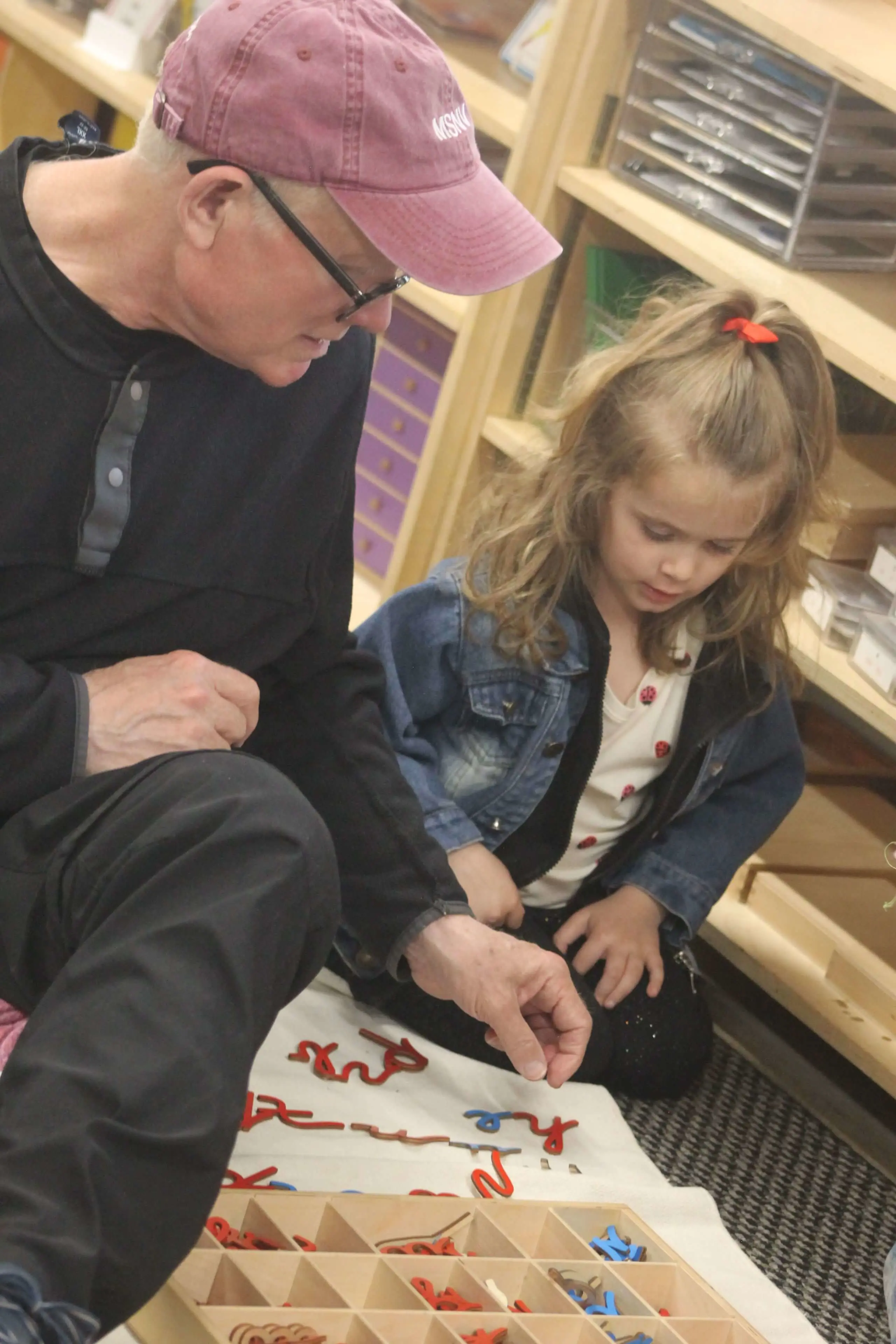  A teacher kneeling beside a primary-age girl working with Montessori letter materials on a classroom rug.