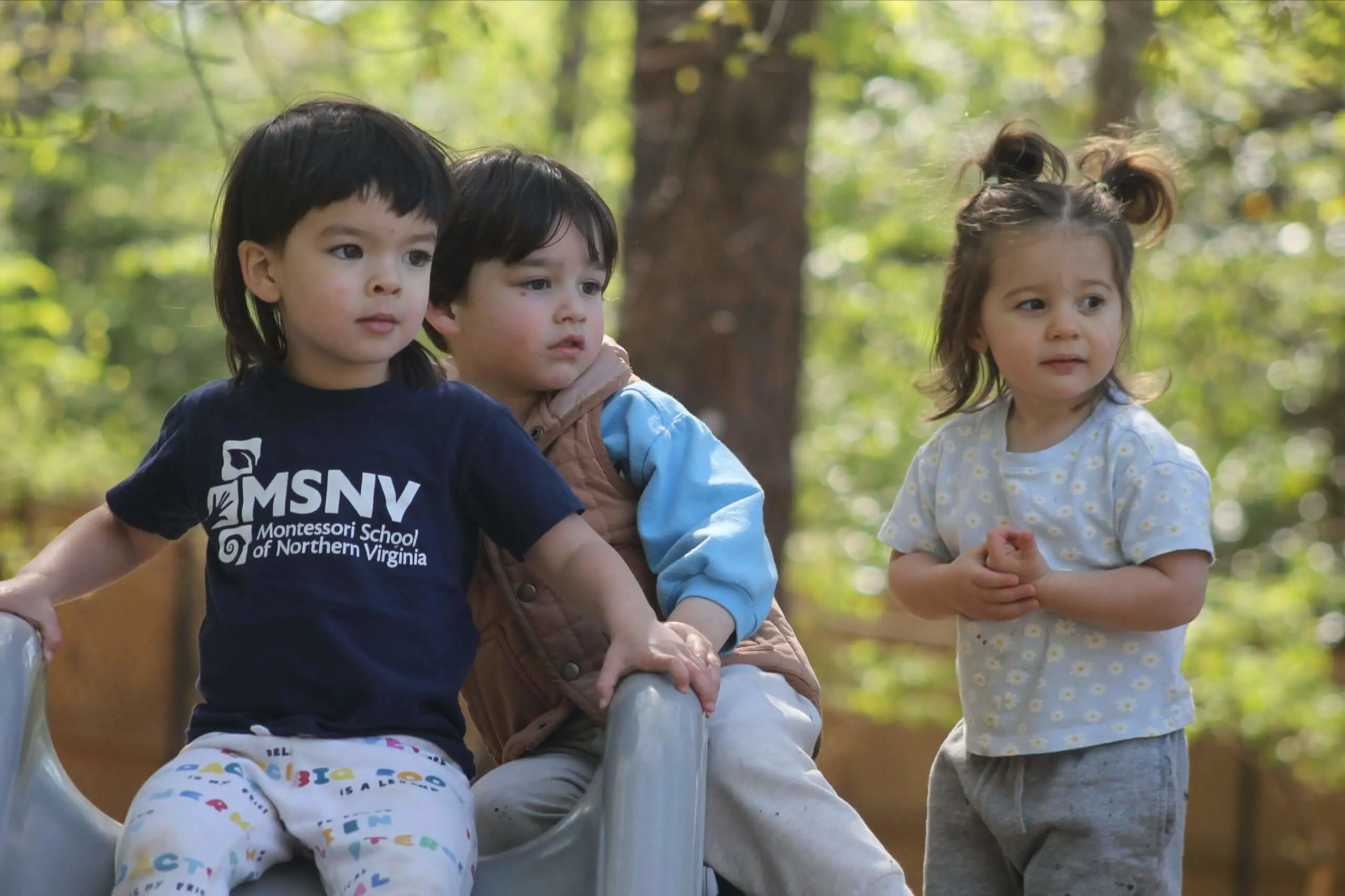 Three MSNV Preprimary students sitting together outdoors, one wearing an MSNV t-shirt.