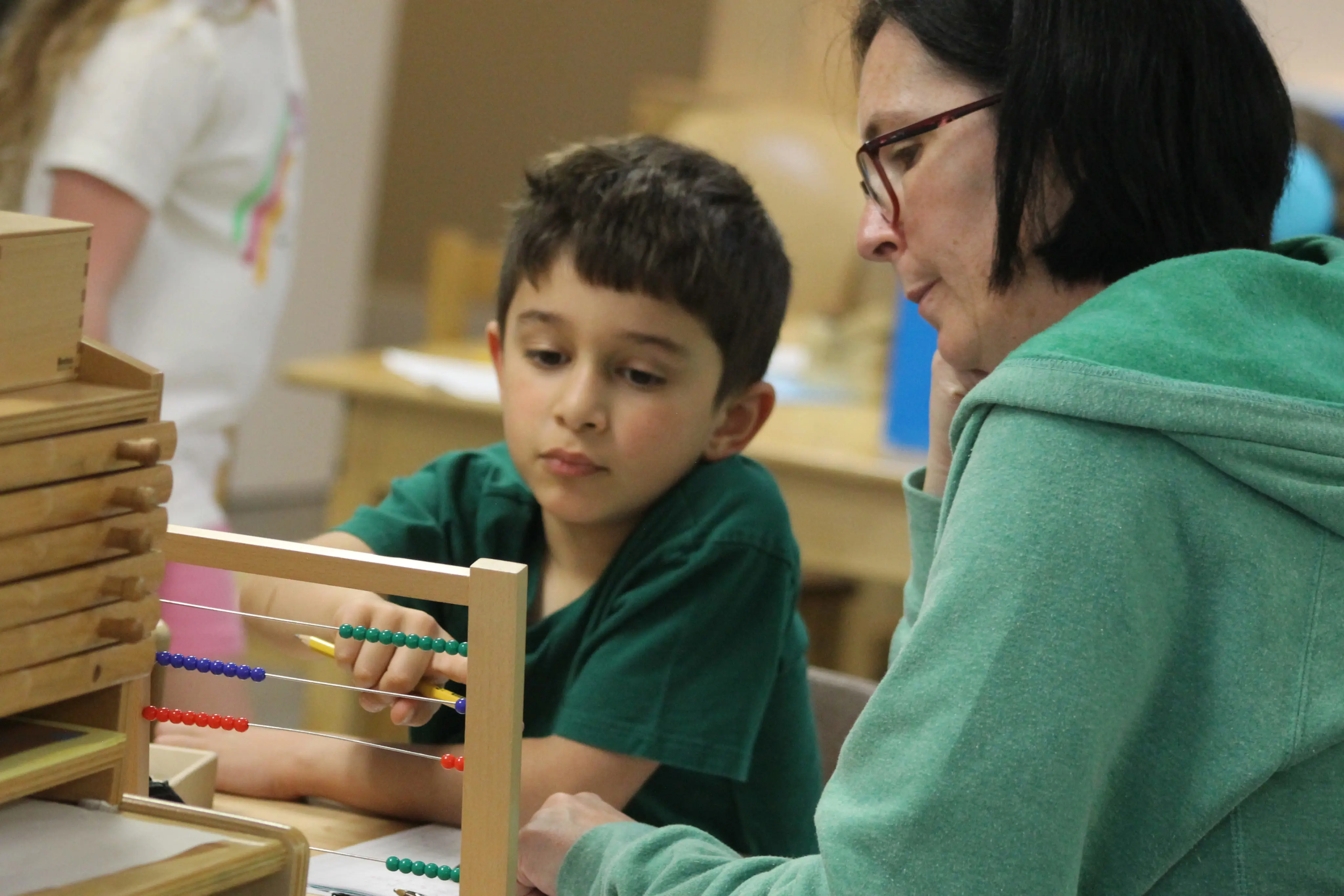 A teacher sitting alongside an elementary boy working with a Montessori bead frame at a classroom table.