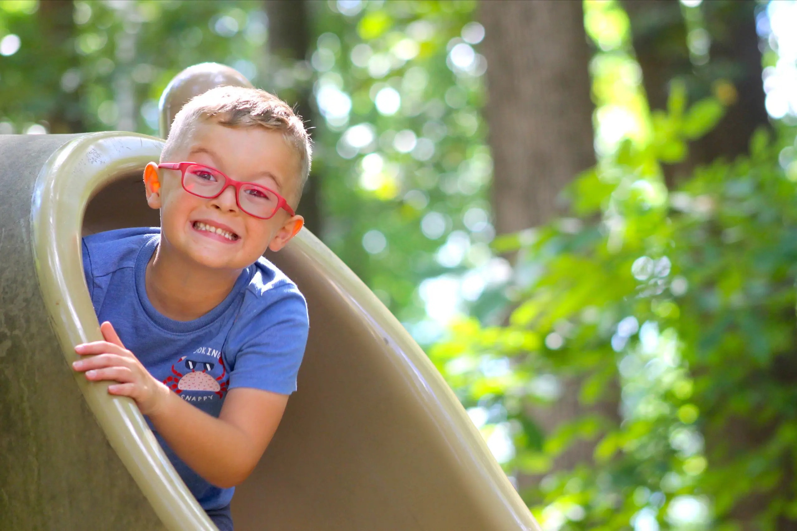 A smiling MSNV primary boy peering through a playground slide outdoors.
