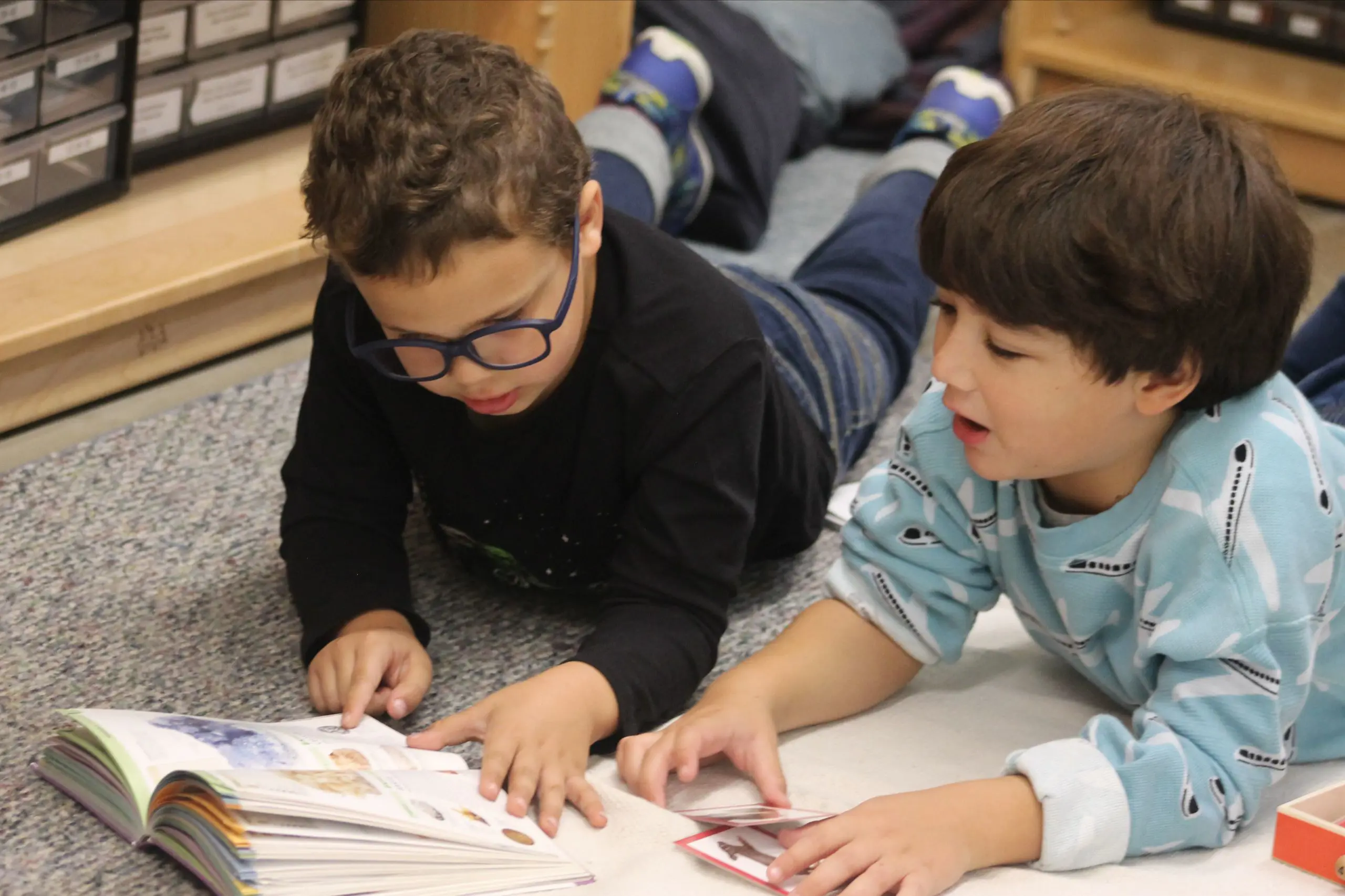 Two MSNV elementary students lying on the floor reading a book together.